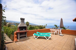 Une terrasse dotée d’une cheminée en pierre, de deux fauteuils, d’une table et d’un parasol.
