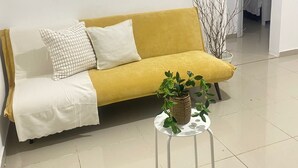 Een gele bank met een witte deken en kussens, een klein wit tafeltje met een plant, en een betegelde vloer.