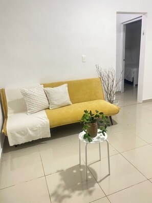 A yellow sofa with a white throw blanket and pillows, a small white table with a plant, and a tiled floor.