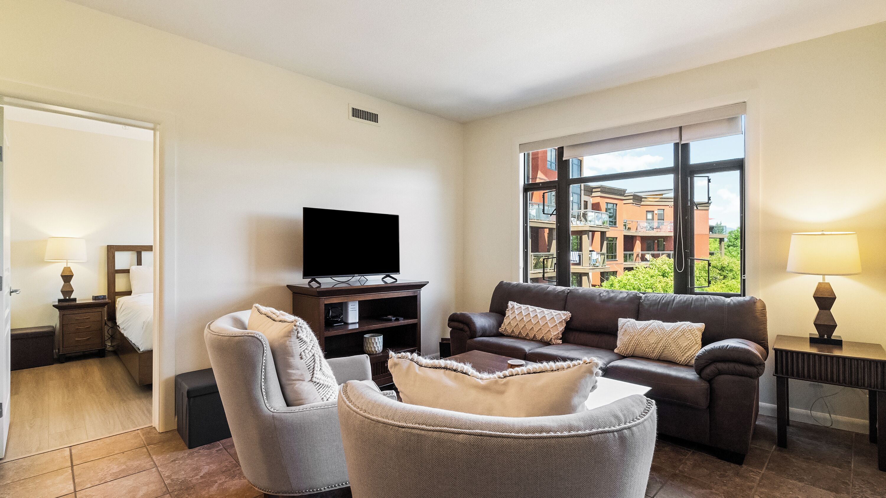 A living room with a sofa, armchair, TV, and a window with a view of buildings.