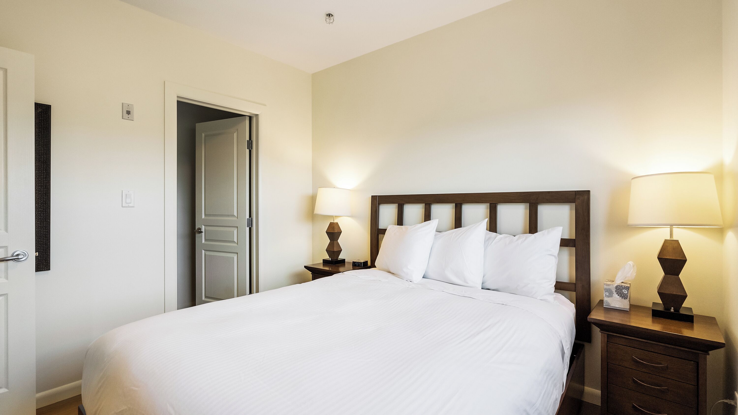 A hotel room with a bed, two bedside tables with lamps, a door, and a wall-mounted light fixture.