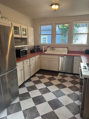 Eine Küche mit Edelstahl-Kühlschrank, Mikrowelle und Geschirrspüler, weißen Schränken, einem Schachbrettfußboden und Fenstern mit Blick nach draußen.