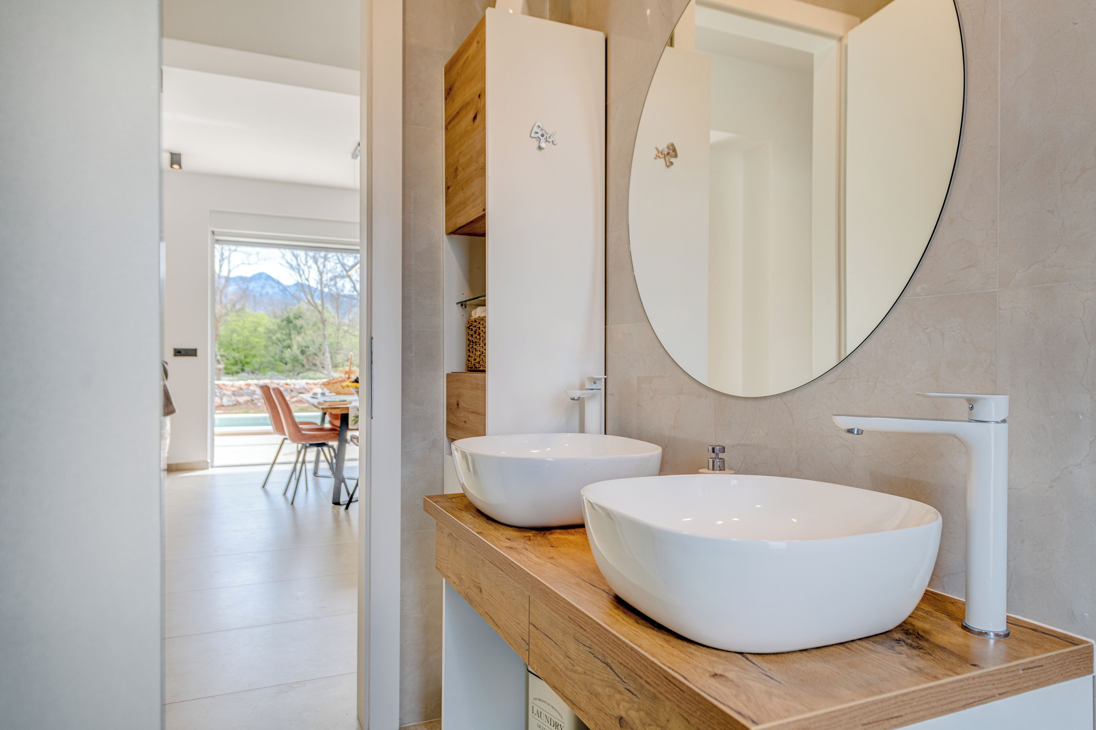 A modern bathroom with two large white sinks, a wooden vanity, a large mirror, and a view of a dining area with a table and chairs.