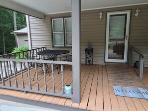 A covered porch with a table and chairs, a welcome mat, and two windows with shutters.