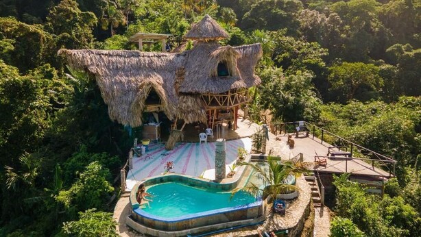 Um edifício com telhado de palha, piscina e palco, cercado por vegetação exuberante.