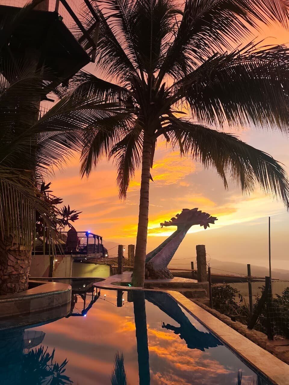 A pool with a sunset view, surrounded by palm trees and a wooden deck.