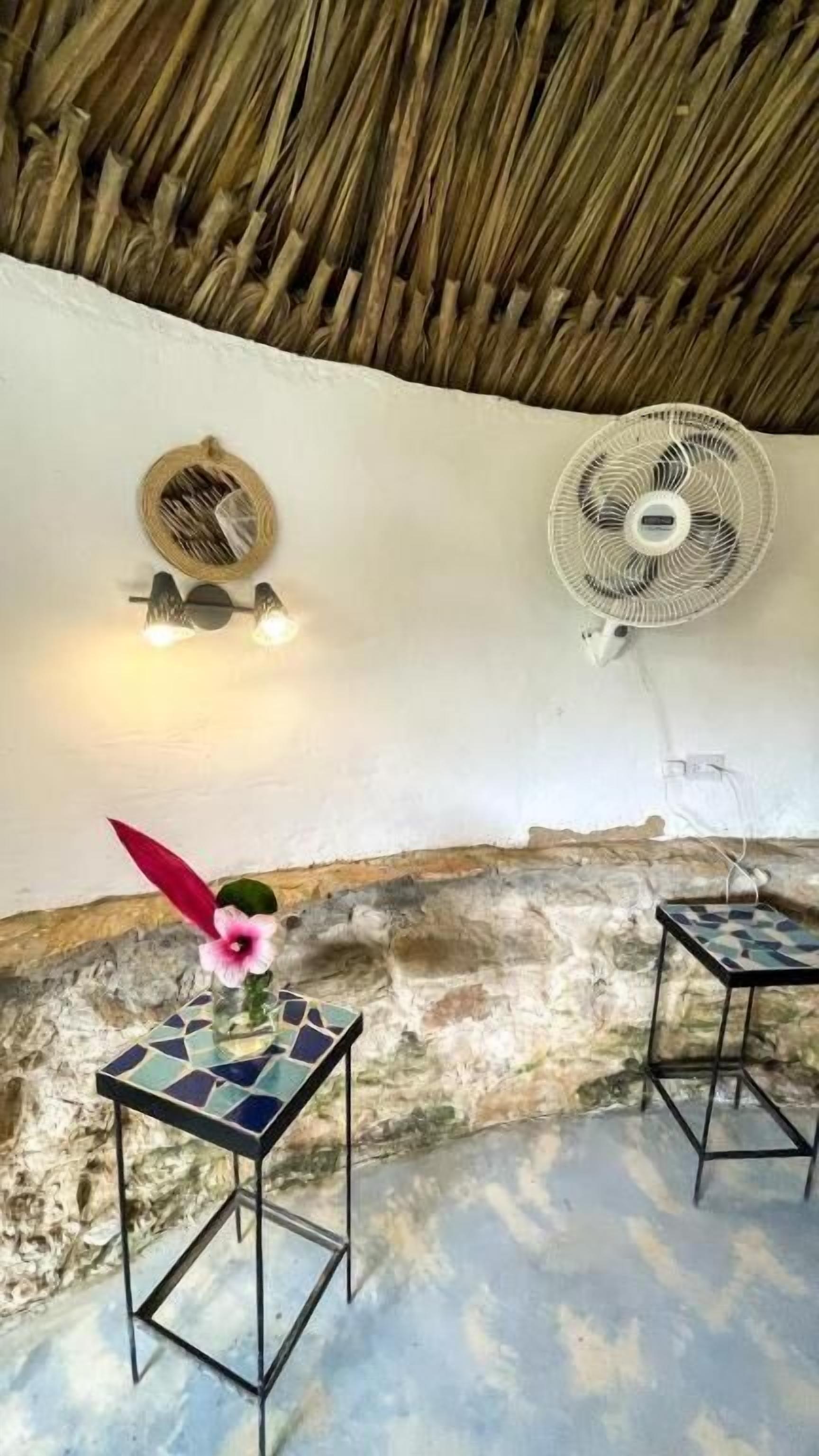 A room with a thatched ceiling, two wall-mounted fans, and two small tables with decorative tiles and a flower.