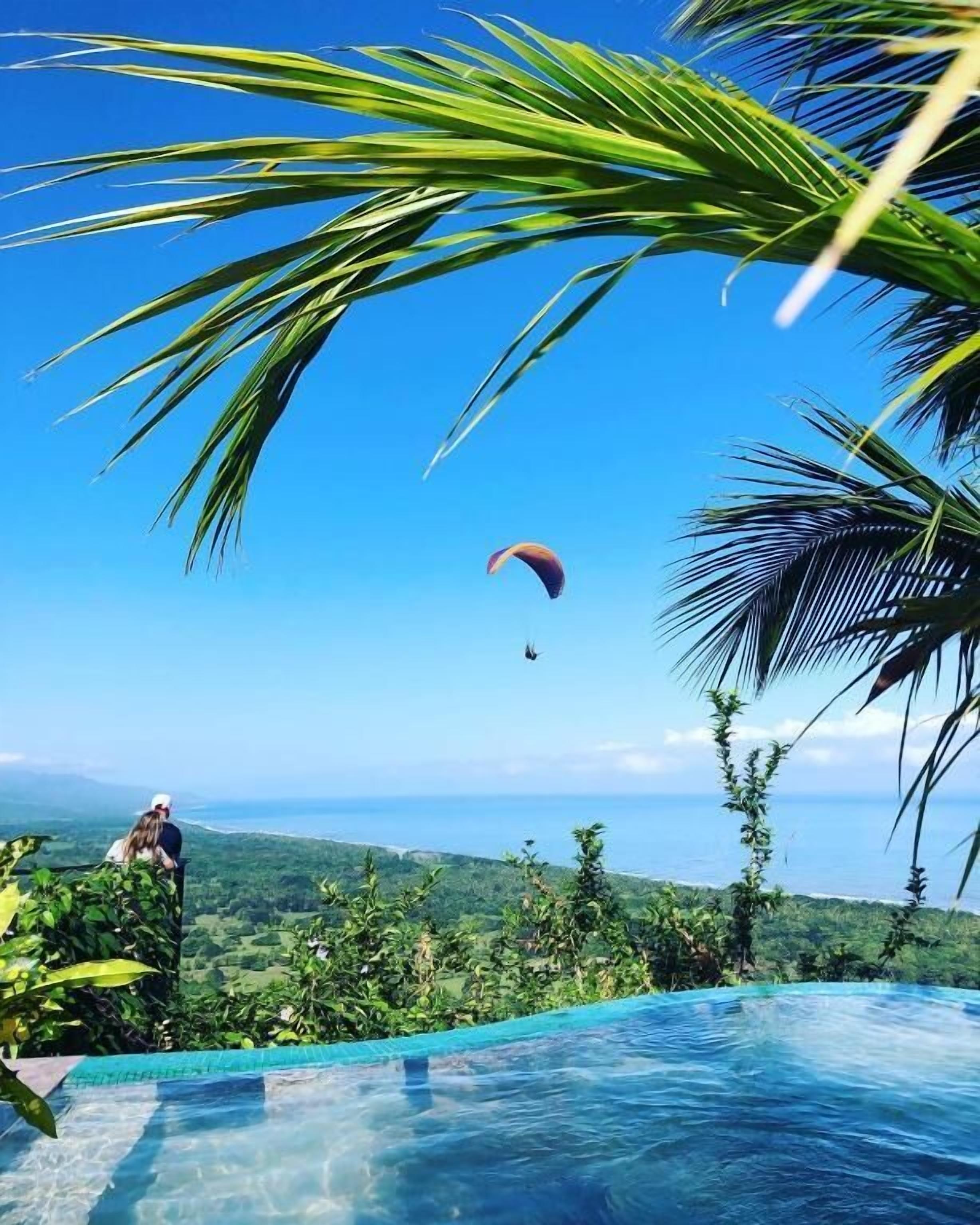 A clear sky, a person on a hill overlooking a pool, and a parachute in the distance.
