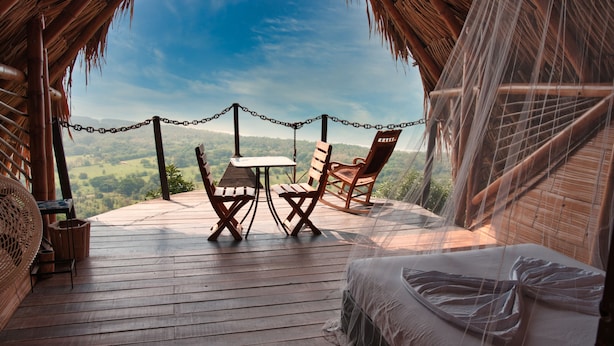 A thatched-roof room with a wooden deck, two wooden chairs, a small table, and a bed with a white sheet.
