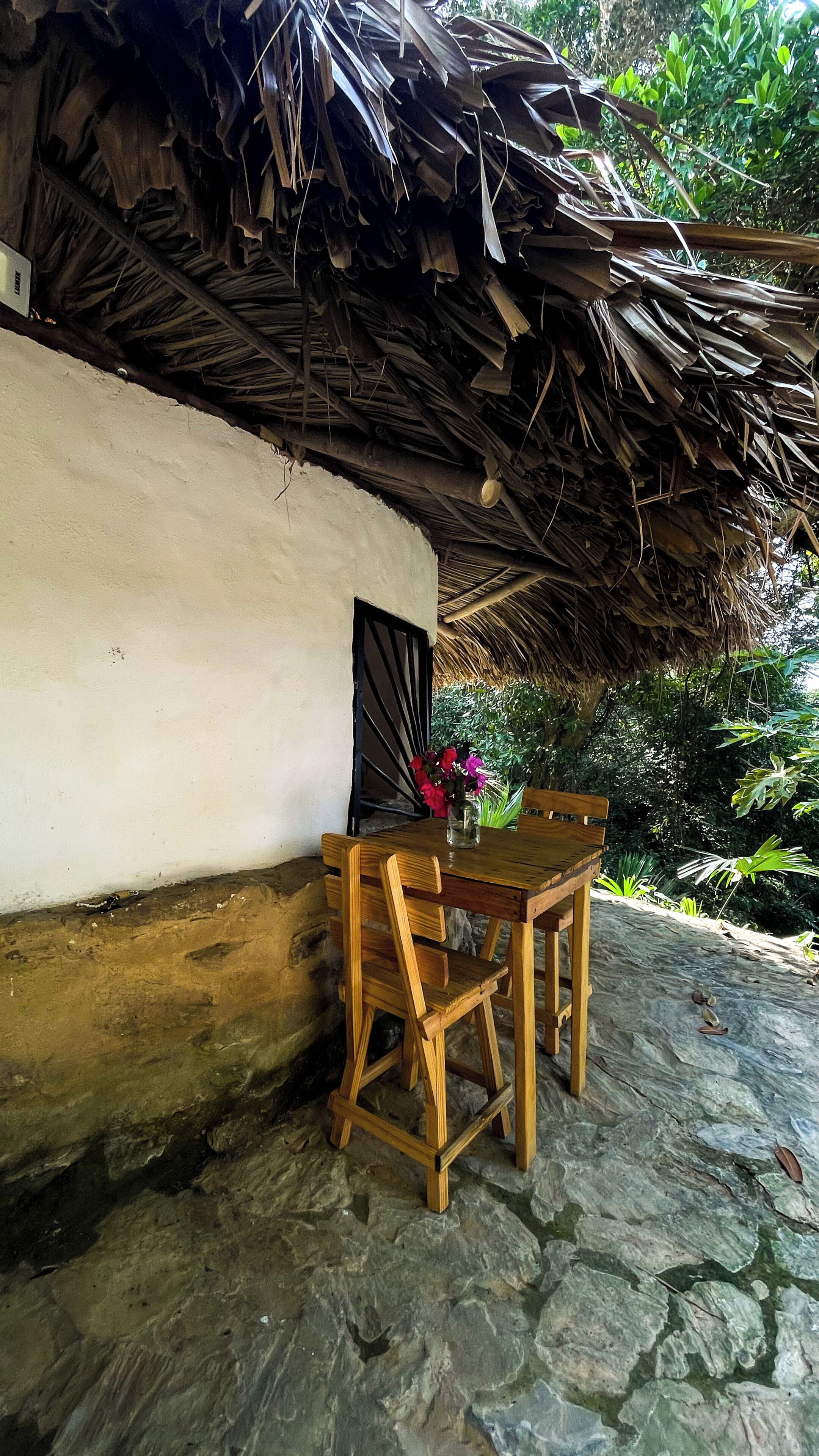 A rustic outdoor seating area with a wooden table and chairs, a thatched roof, and a stone floor.