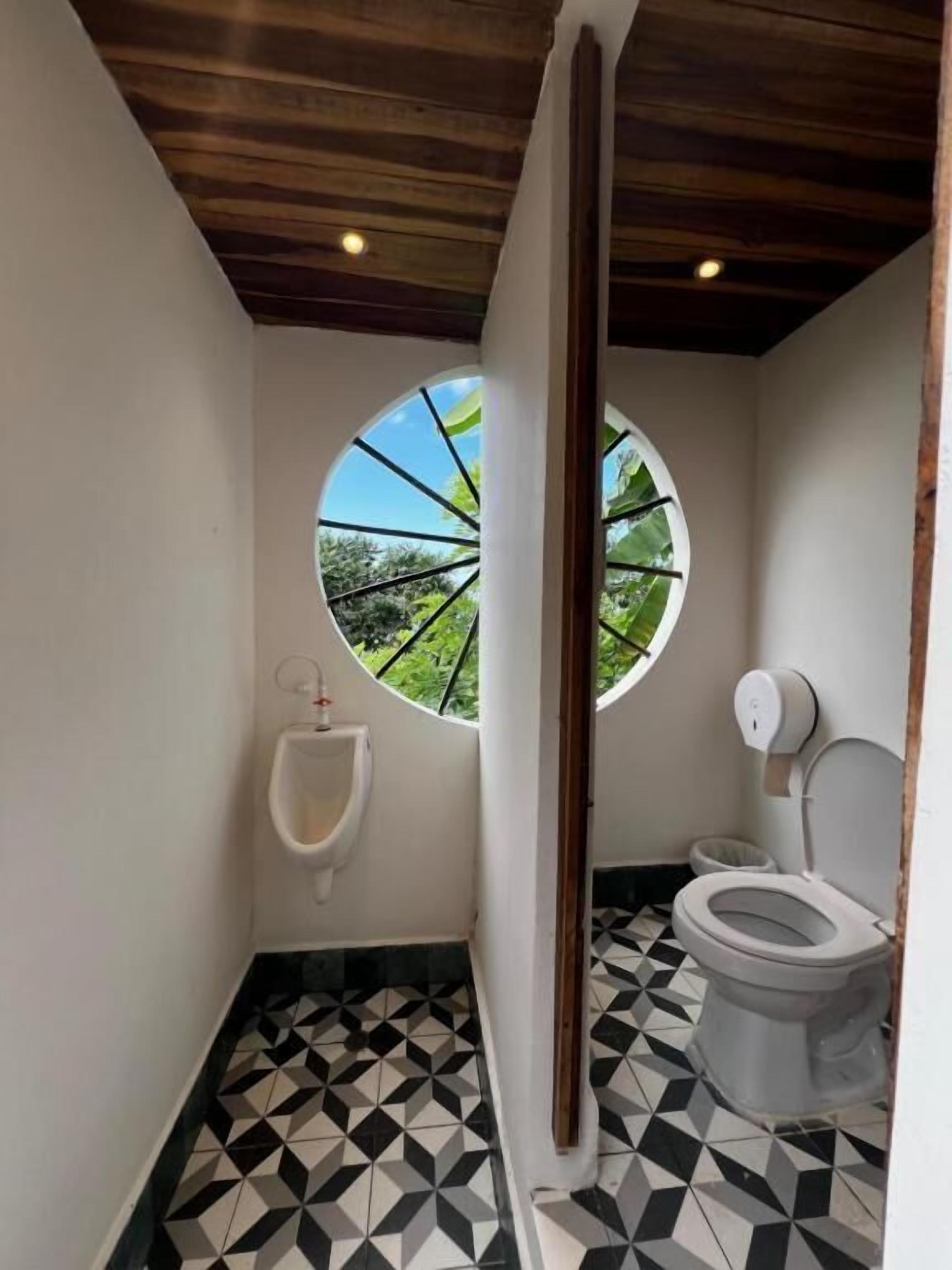 A bathroom with a urinal, toilet, and a circular window with a view of greenery.