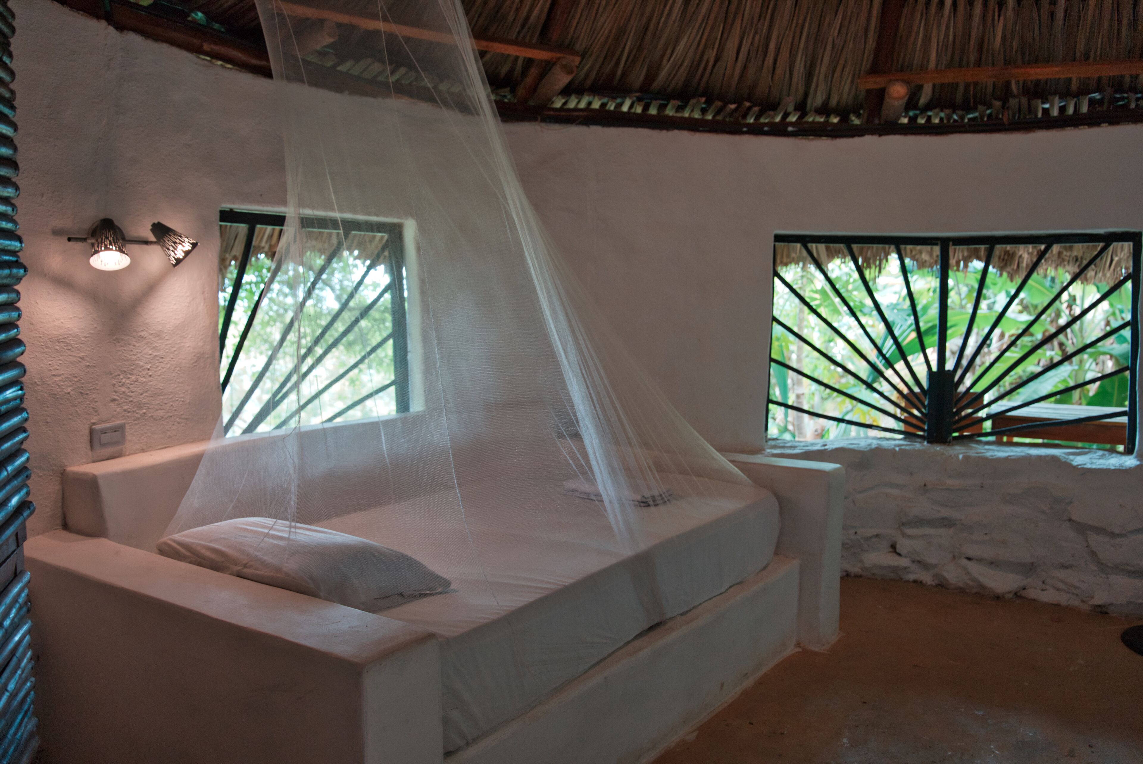 Une chambre avec un lit sous une moustiquaire, une fenêtre donnant sur un paysage verdoyant et un toit de chaume.