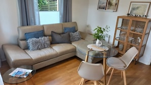 Un salon avec un canapé beige, une table basse en verre, deux fauteuils, une petite table ronde ornée d’une plante et un meuble avec des portes en verre.