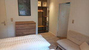 Une chambre à coucher comprenant un lit, une commode en bois, une armoire avec des vêtements suspendus et une porte donnant sur une autre pièce.