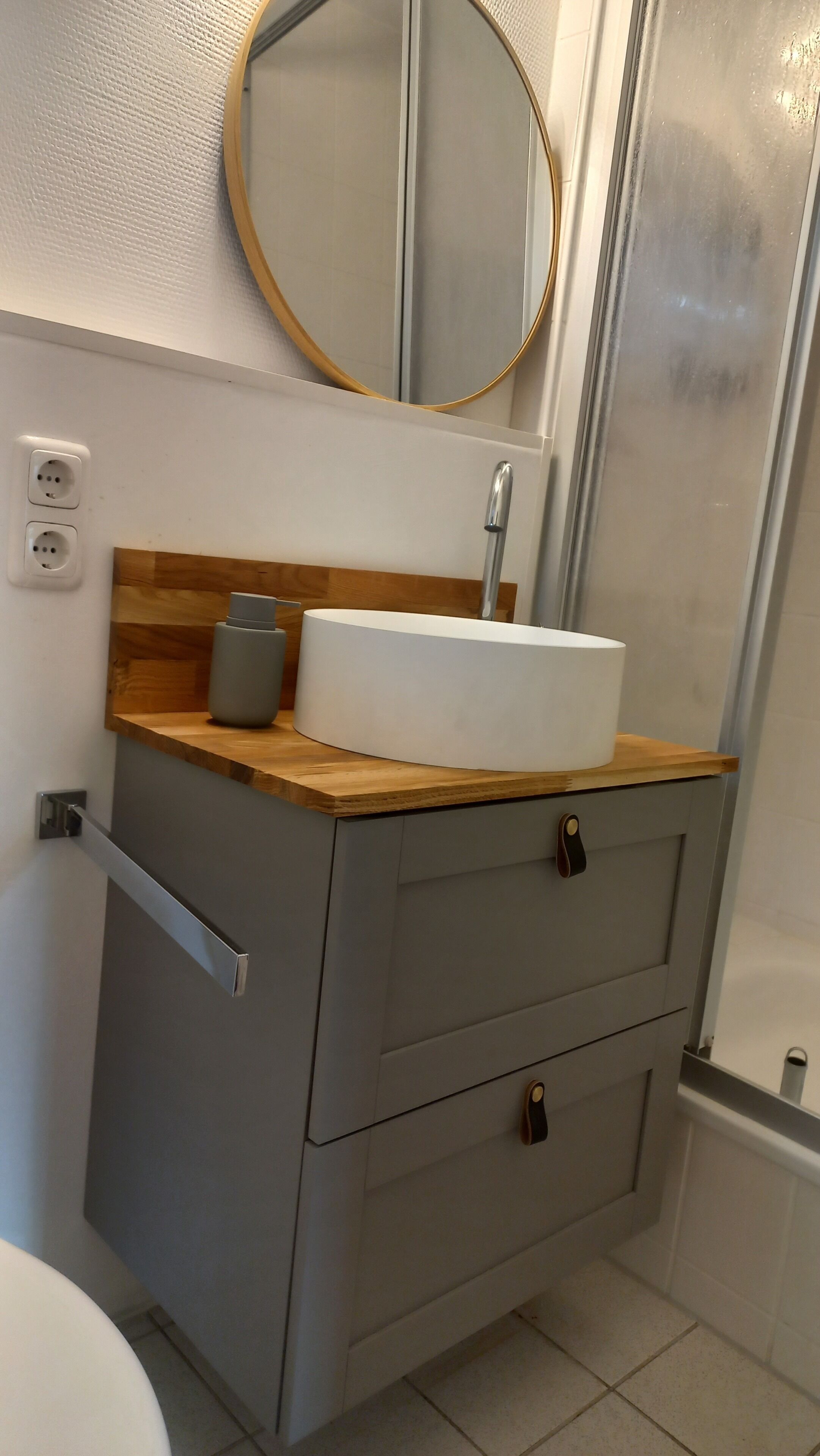 A bathroom with a white sink, wooden countertop, grey cabinet, and a round mirror.