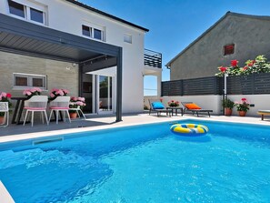 Ein moderner Poolbereich im Freien mit überdachter Sitzecke, Liegestühlen und Topfpflanzen.