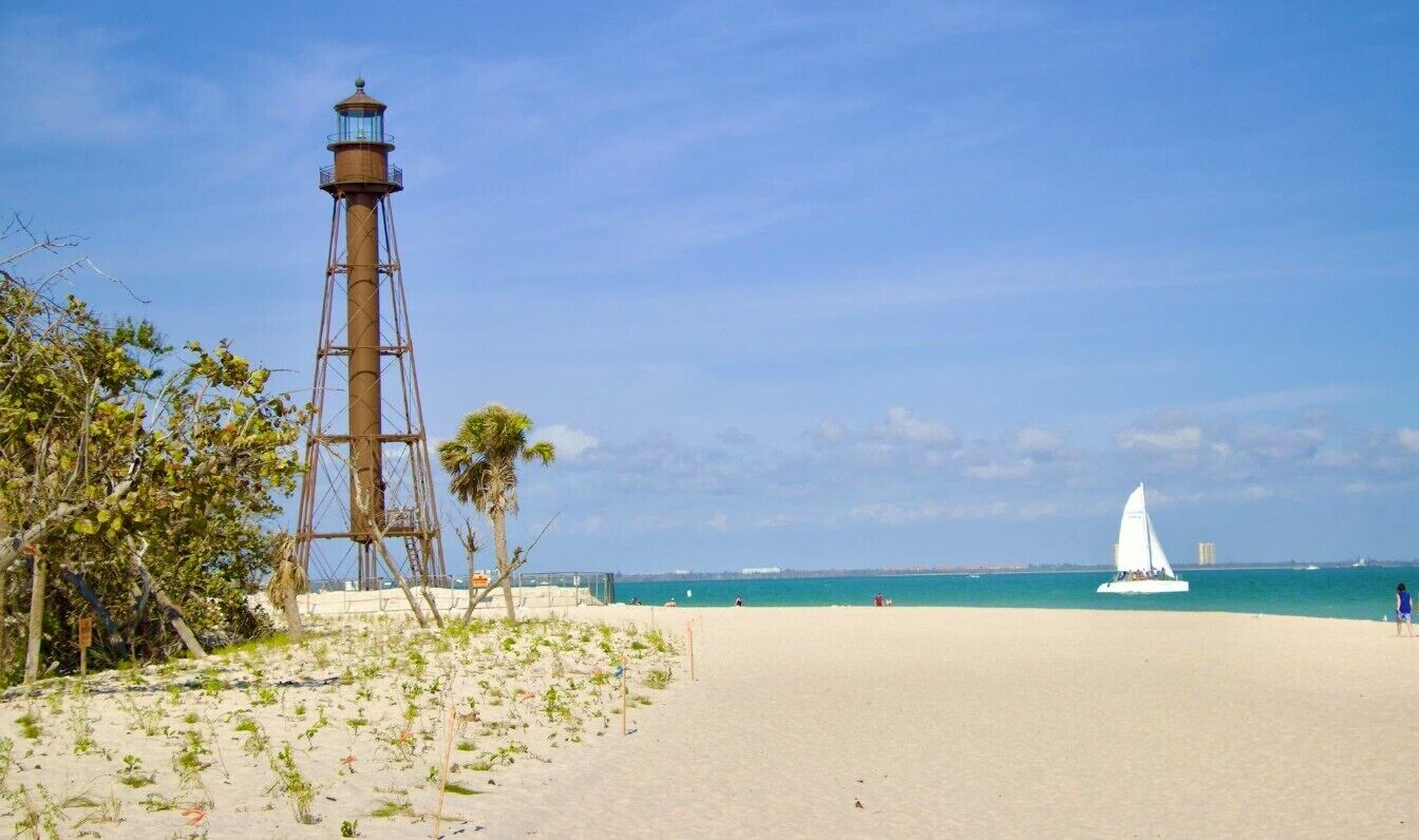 A beach with a lighthouse, clear sky, and a sailboat on the water.