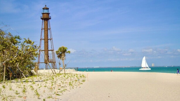 A beach with a lighthouse, clear sky, and a sailboat on the water.