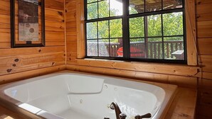Ein Holzblockhaus-Interieur mit einer großen Badewanne, einem Fenster mit Blick ins Grüne und einem gerahmten Bild an der Wand.