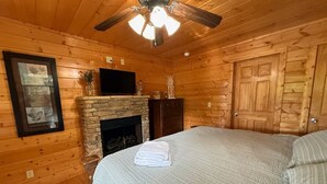 Ein Holzhäuschen-Zimmer mit Kamin aus Stein, einem Bett, einem Fernseher und einem Deckenventilator.