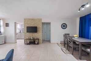 A spacious living area with a flat-screen TV mounted on a stone wall, a dining table with chairs, and a kitchen in the background. - Cozy 3BR Condo Pool  10 minutes to Airport (Carolina)