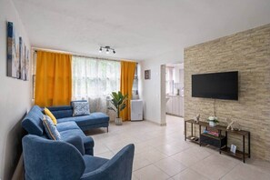 A living room with a sofa, a TV mounted on a brick wall, and a kitchen in the background. - Cozy 3BR Condo Pool  10 minutes to Airport (Carolina)