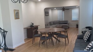 A modern living room with a dining area, grey sofas, a chandelier, and a ceiling fan.