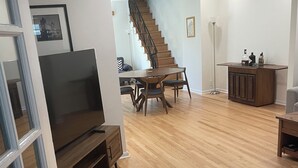 A living room with a wooden floor, a dining table with chairs, a TV on a stand, a staircase, and a small cabinet.