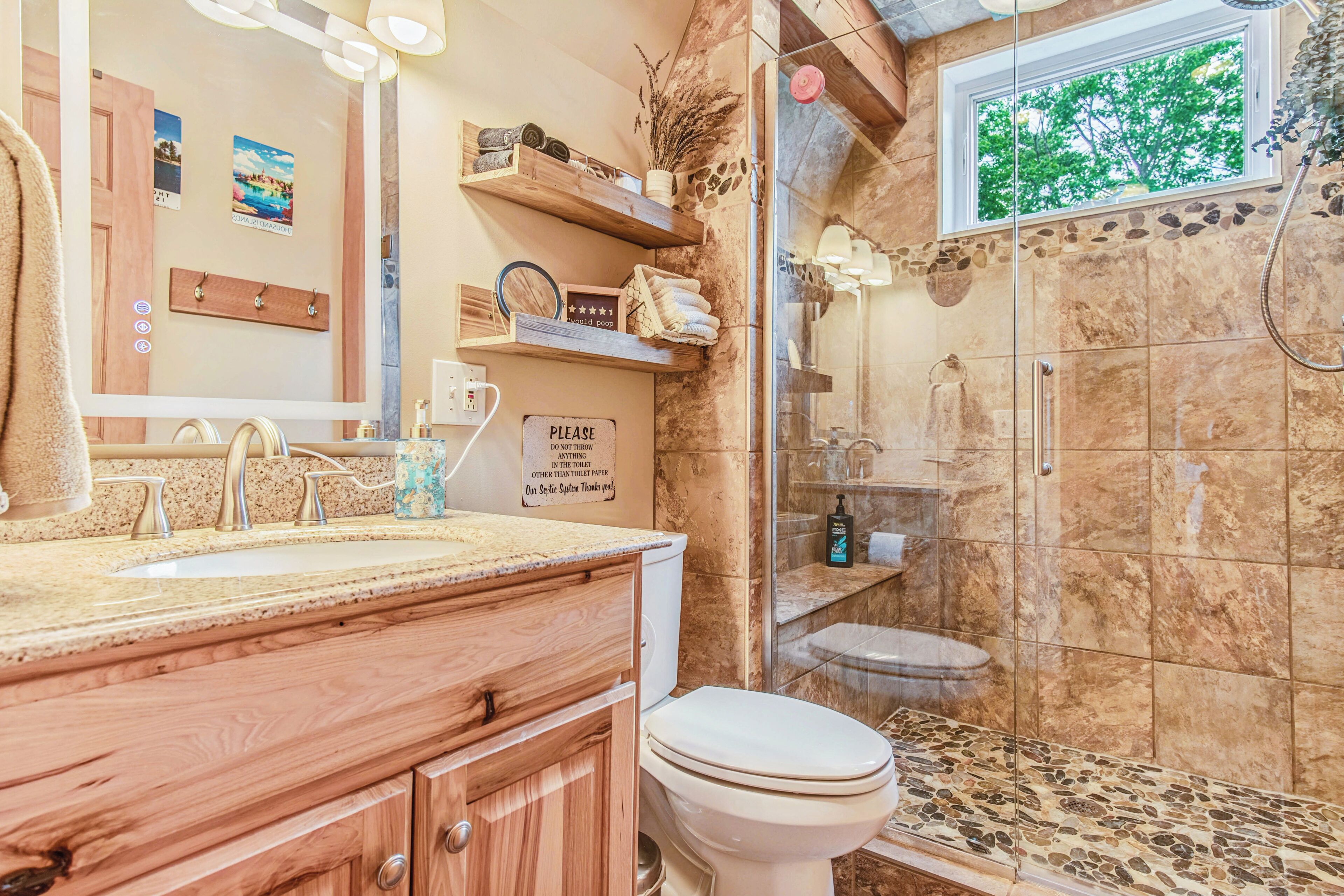 Une salle de bain avec un meuble-lavabo en bois, des toilettes, une douche avec une porte en verre et une fenêtre donnant sur les arbres.