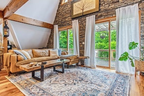 Una habitación tipo loft acogedora con pared de piedra, techo de madera, un amplio ventanal con cortinas, un sofá, una mesa de centro y una alfombra estampada.
