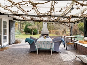 Eine Terrassengruppe im Freien mit Korbmöbeln, einem Tisch mit einer blumigen Tischdecke und einem Kamin aus Ziegelsteinen.
