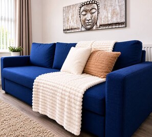 A blue sofa with a white and brown throw blanket, a decorative Buddha painting, and a potted plant. - Walk to Anfield | Sleeps 6 | 2 Beds + Sofa Bed (Liverpool)