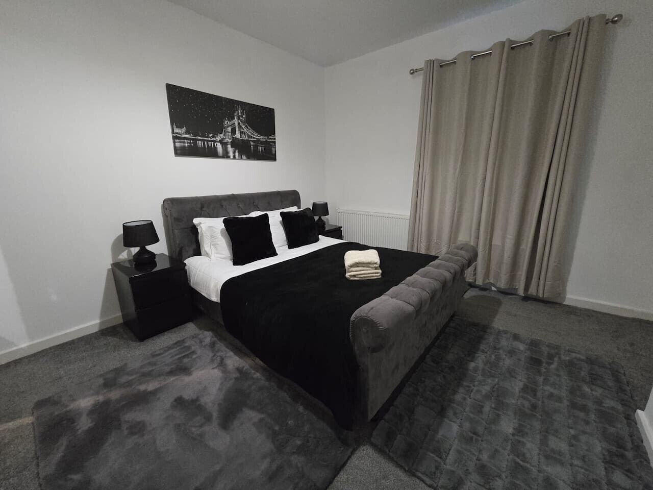 Ein Schlafzimmer mit einem Bett, Nachttischen, einer Lampe, einem Fenster mit Vorhängen und einem Wandbild.