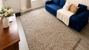 Ein Wohnzimmer mit blauem Sofa, einem beigen Teppich, einem Holztisch, einer weißen Vase mit Blumen, einem an der Wand hängenden Kunstwerk und einem Fenster mit Vorhängen.