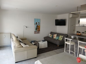 A modern living room with a sofa, a coffee table, a TV, and a kitchen area with a bar. - Beautiful 3-bedroom apartment (70 square meters) in Juan (Antibes)