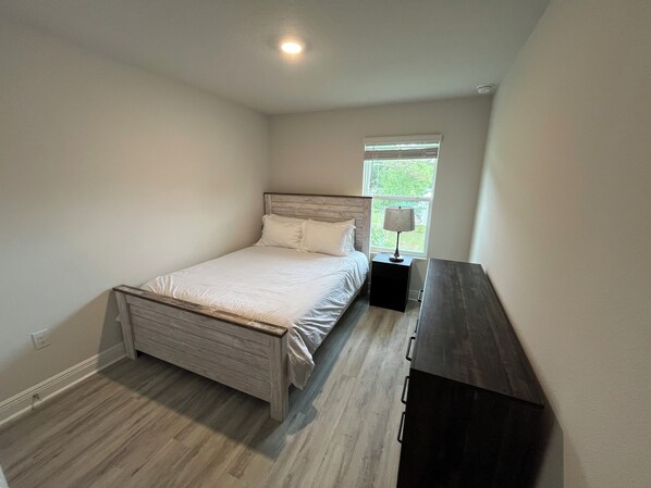 A bedroom with a bed, a nightstand, a dresser, and a window with blinds. - Meadow Flower Townhome-Downtown, Beaches, & UWF (Pensacola)