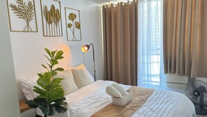 Un lit bien fait, recouvert d’une couvre-lit blanche et beige, avec une plante sur la table de chevet et des œuvres d’art accrochées au mur.