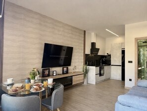 Ett modernt vardagsrum med en platt-TV, ett matbord med frukosttillbehör, en köksdel med en spis och en grå soffa.