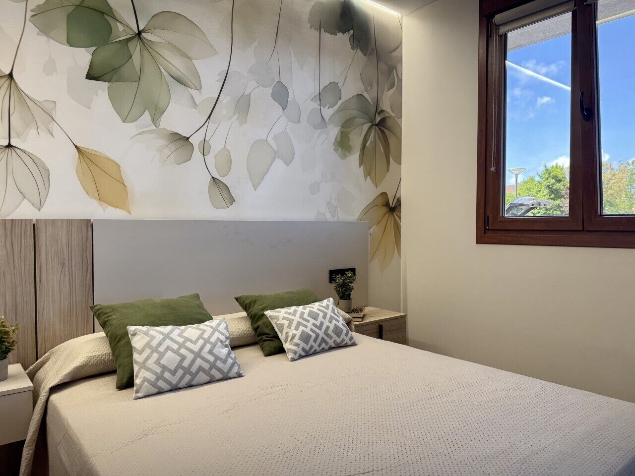 A bedroom with a bed, pillows, a nightstand, a window, and a wall with a leaf pattern.