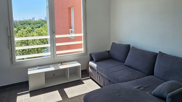 Una sala de estar moderna con un sofá seccional, un ventanal con vistas a zonas verdes y edificios, y una lámpara de techo.