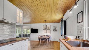 Moderni keittiö, jossa on puinen katto, valkoiset kaapit, ruokailutila pöytineen ja tuoleineen sekä suuri ikkuna.