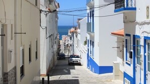 Une rue étroite pavée, bordée d’immeubles, un ciel d’un bleu limpide et une vue lointaine sur la mer.