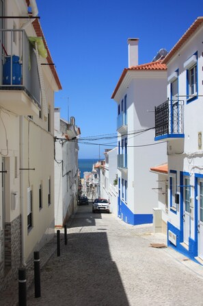 A narrow cobblestone street lined with buildings, a clear blue sky, and a distant view of the sea. - Rustic house featuring traditional Portuguese handicrafts (Nazaré)