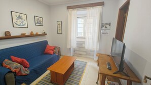Un salon avec un canapé bleu, une table basse en bois, un téléviseur posé sur un meuble et une fenêtre avec des rideaux.