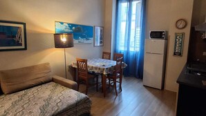 Ein kleiner, gemütlicher Wohnraum mit einer Couch, einem Esstisch mit Stühlen, einer Küchenzeile mit Kühlschrank und einem Fenster mit Vorhängen.