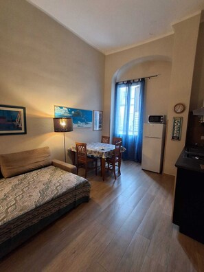 A small, cozy living room with a sofa, a dining table with chairs, a kitchen area with a refrigerator, and a window with curtains. - Apartment for families and groups of friends in the heart of Naples (napoli)