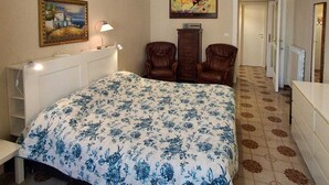 Ein Schlafzimmer mit einem Bett, zwei Stühlen, einer Kommode, einem Gemälde und einem Flur.