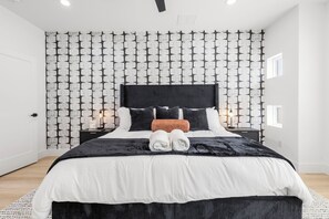 Et moderne hotelværelse med tapet med mønster, en sort sengegavl, en pænt redt seng med sort og hvidt sengetøj og et lille natbord med en lampe.