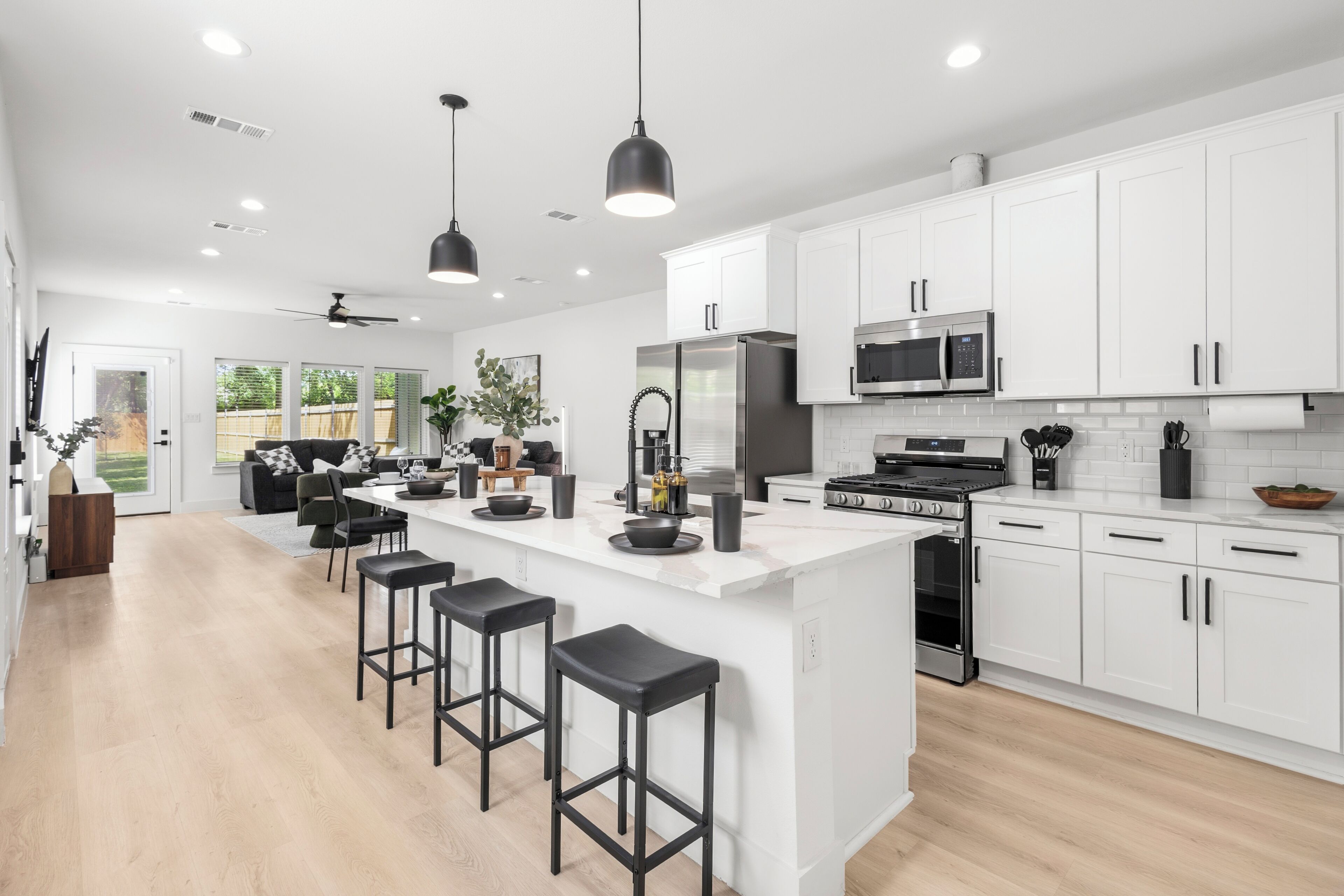 Una cocina moderna con armarios blancos, una isla central con taburetes, electrodomésticos de acero inoxidable e iluminación colgante.