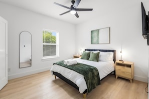 Ein Schlafzimmer mit Bett, Deckenventilator, Nachttisch, Fenster mit Jalousien und Wand-Fernseher.